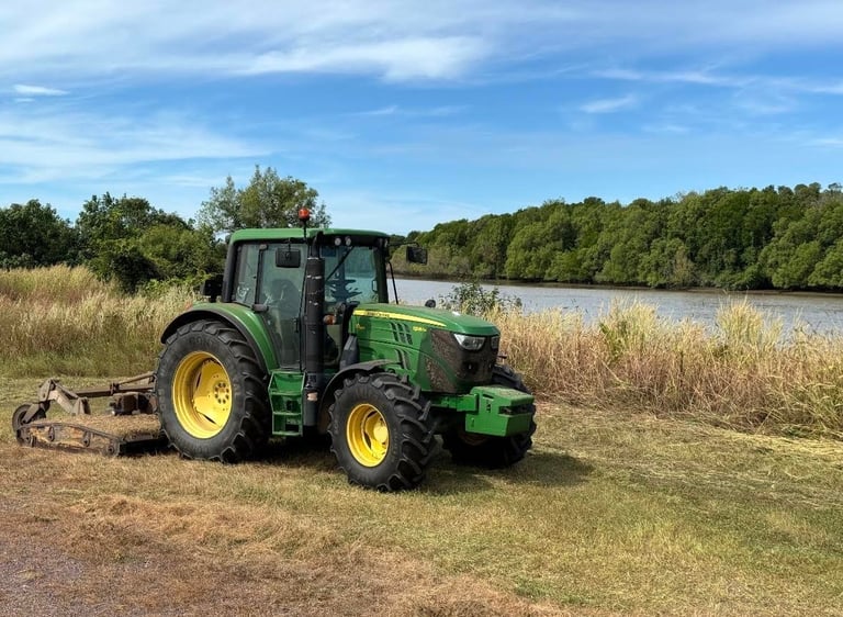 Green and yellow John Deere tractor with attached mower in a grassy field near a lake and wooded area