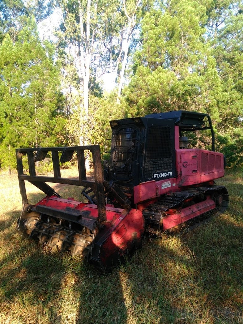 Red and black tracked forestry equipment with a loading grapple attachment parked in a grassy area under tall eucalyptus trees