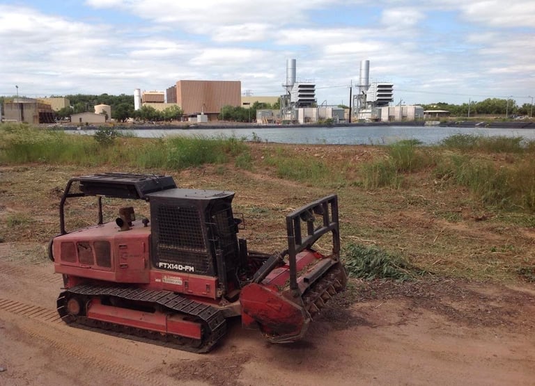 Red tracked vehicle with excavator bucket parked on dirt ground near waterfront industrial buildings